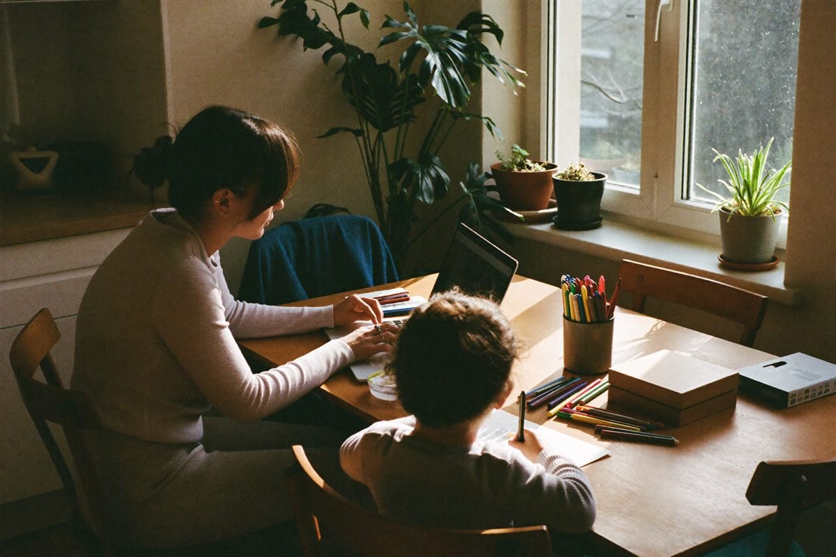 Mama dirba nešiojamu kompiuteriu prie virtuvės stalo, šalia vaikas piešia flomasteriais